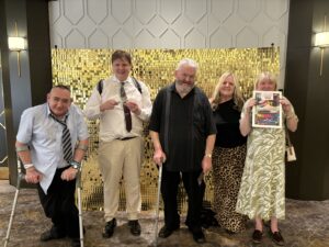 Five people stand smiling in front of a gold sequin backdrop at the TILII Roving Report: A Visit to the University of Atypical for Arts and Disability, celebrating with a “WINNER” certificate and dressed in semi-formal attire.
