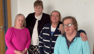 Four women stand together indoors, posing for a group photo against a plain white wall. Dressed in casual clothing, they smile warmly, embodying the spirit of TILII: Talking About Mental Health and supporting one another.