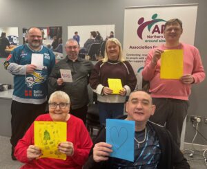 Six adults smiling and holding up handmade cards with hearts and festive drawings, posing in a community center in front of a Carers Rights Day banner for the Association for Independent Living (A.I.L.).