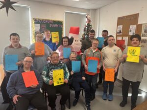 A group of people are smiling and holding colorful hand-made cards in a festive room decorated with tinsel, a bulletin board, and a Christmas tree with a Santa figure, celebrating Safeguarding Week 2025 together.