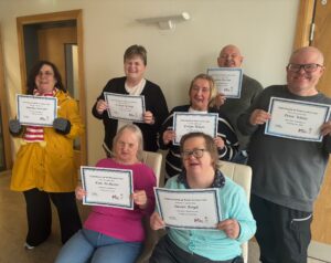 Seven smiling adults pose indoors, each holding a certificate of participation for Carers Rights Day. They appear happy and proud, seated and standing in two rows against a neutral-colored wall with a wooden door visible on the left.