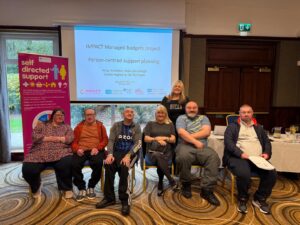 Seven people are sitting and standing in front of a projection screen displaying "IMPACT Managed budgets project: Person-centred support planning." Celebrating Human Rights Day 2025, they smile at the camera beside a "self-directed support" banner.