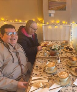 Four adults sit around a small café table with coffee, tea, and cake. Warm lights are strung along the wall for a cozy atmosphere as they chat and enjoy desserts, celebrating inclusion on International Day of Persons with Disabilities 2025.