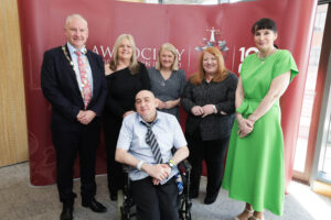 Six adults, including one man in a wheelchair, pose together in front of a red backdrop with white text and a crest. Celebrating the spirit of Living Well, Living Longer, four women wear dresses—one in green—and two men wear suits.