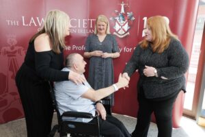 Four people are gathered, with one in a wheelchair shaking hands with a woman. Two other women stand nearby, smiling—a warm display of community spirit. Behind them is a red banner for the Law Society of Northern Ireland, emphasising the importance of Living Longer, Living Well within a supportive environment.